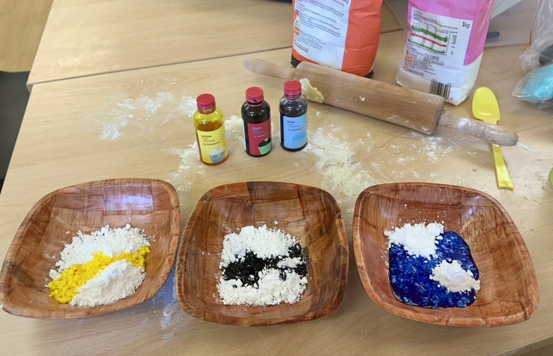 Three wooden bowls of icing sugar being mixed with yellow, black and blue food colouring