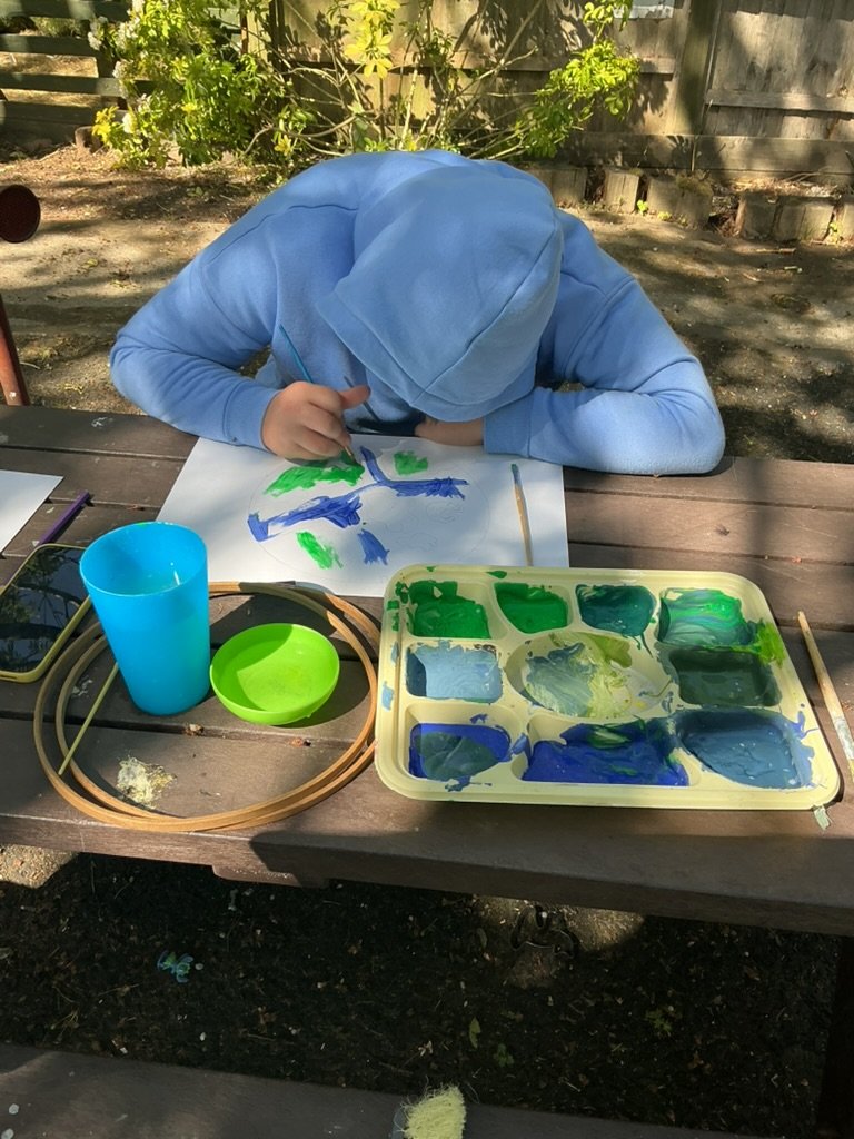 Child painting outdoors at a picnic table with a tray of green and blue paints