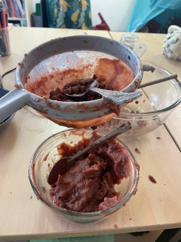 Red fruit pulp being pushed through a sieve into a glass bowl
