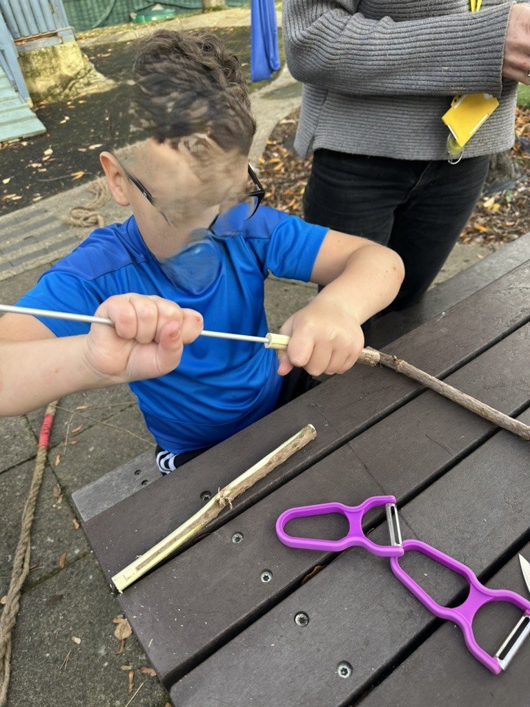 Child working on a stick with a peeler at an outdoor picnic table