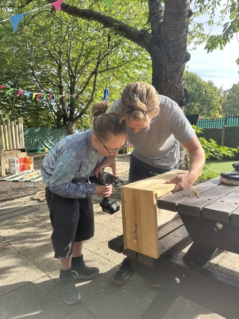 Child using a cordless drill to build a wooden box, with an adult helping